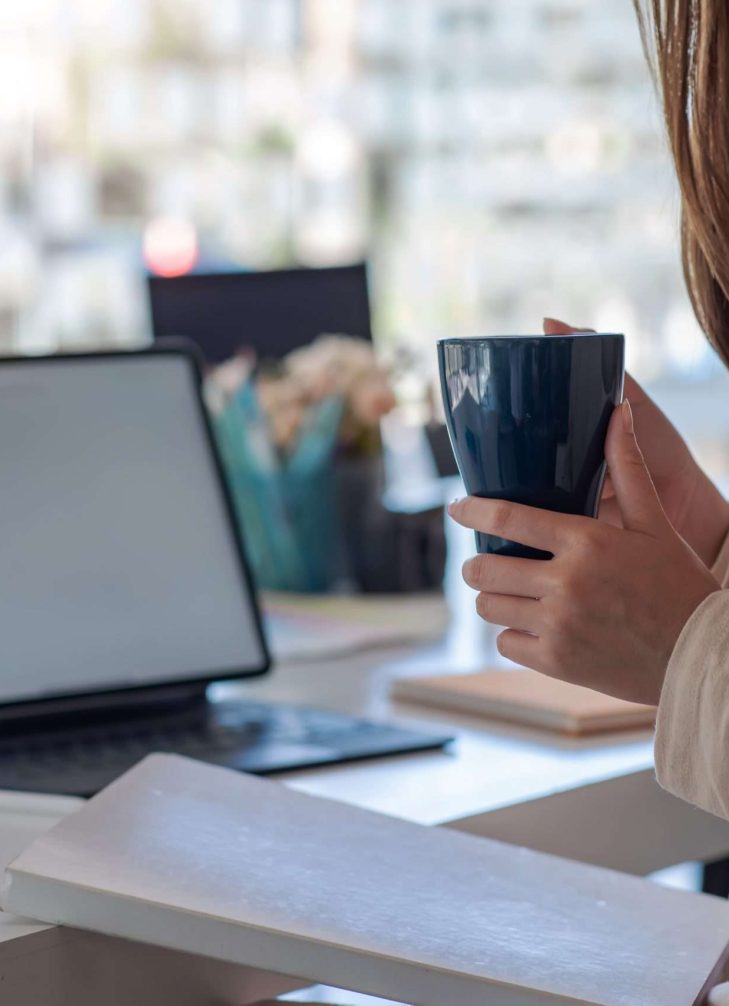 Image of a woman holding a coffee and a tablet on the desk in her office.